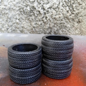 May include: Four black model car tires stacked in pairs. The tires have a detailed tread pattern and a circular shape. The background is a textured gray wall and a red surface.