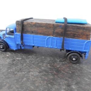 Op de afbeelding: Een blauwe speelgoedtruck met een houten kist en een blauwe zeil op de bovenkant. De truck heeft zwarte banden en een zwart touw dat de kist op zijn plaats houdt.