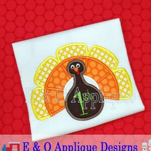 May include: A white t-shirt with an embroidered turkey applique. The turkey is brown with a yellow and orange fan-shaped tail. The turkey has a brown head with a red wattle and a yellow beak. The turkey has a brown body with a green stem on top. The t-shirt has the text "E & O Applique Designs" printed on the bottom.