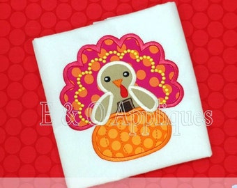 May include: A white t-shirt featuring an applique of a pink and orange turkey holding a pumpkin. The turkey has a brown beak and brown eyes. The text "E & O Appliques" is visible in the background.