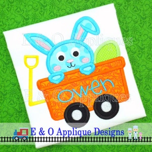 May include: A blue bunny with pink ears and white spots is sitting in an orange wagon with black wheels. The wagon has the name "Owen" written on the side. The bunny is holding a green egg in its paws.