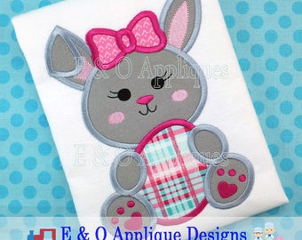 May include: A gray bunny with pink ears and a pink bow holding a plaid heart. The bunny is embroidered on a white t-shirt.