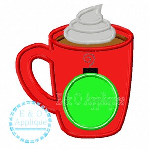 May include: Red mug with a green bauble hanging from the handle. The mug is filled with brown liquid and topped with white whipped cream.