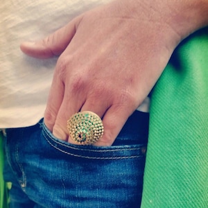 Pu&ograve; includere: Un anello d'oro con una grande pietra preziosa verde al centro. L'anello &egrave; incastonato con pietre preziose verdi pi&ugrave; piccole intorno alla pietra centrale.