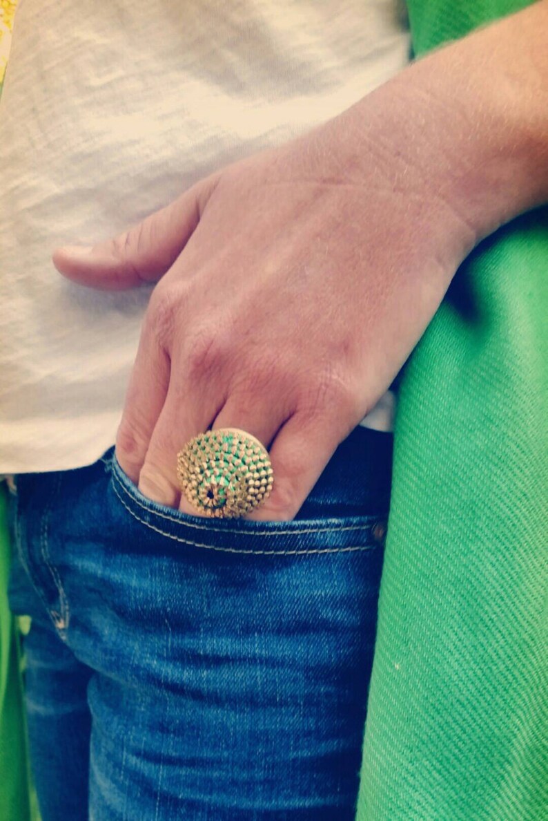 Pu&ograve; includere: Un anello d'oro con una grande pietra preziosa verde al centro. L'anello &egrave; incastonato con pietre preziose verdi pi&ugrave; piccole intorno alla pietra centrale.