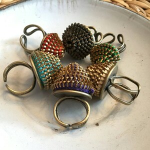 Pu&ograve; includere: Sei anelli colorati con un design unico. Ogni anello presenta una parte superiore rotonda e testurizzata realizzata in metallo e tessuto. Gli anelli sono disposti in cerchio su una superficie bianca.