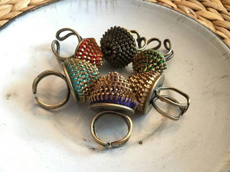 Pu&ograve; includere: Sei anelli colorati con un design unico. Ogni anello presenta una parte superiore rotonda e testurizzata realizzata in metallo e tessuto. Gli anelli sono disposti in cerchio su una superficie bianca.