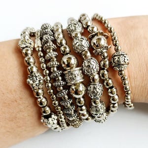 Pode incluir: Um conjunto de várias pulseiras de contas em tom prateado usadas juntas. As pulseiras apresentam uma variedade de formas de contas metálicas, incluindo esferas lisas, padrões florais texturizados e designs intrincados de filigrana.