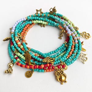 Pode incluir: Uma coleção de pulseiras de contas em várias cores, incluindo turquesa, laranja, vermelho e roxo. Cada pulseira é adornada com pingentes dourados, como corujas, borboletas e rostos sorridentes. As pulseiras estão dispostas em um padrão circular.
