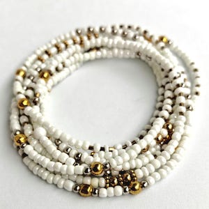 Pode incluir: Uma coleção de contas de cintura brancas com detalhes dourados e prateados. As contas são dispostas em vários fios, criando um efeito em camadas. As contas douradas são maiores e mais proeminentes.