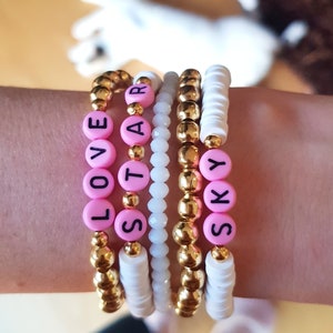 Puede incluir: Un conjunto de seis pulseras con cuentas rosas y cuentas doradas. Las cuentas están escritas con letras para deletrear las palabras "LOVE", "STAR" y "SKY".
