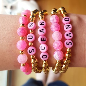 Pode incluir: Quatro pulseiras de contas cor-de-rosa com espaçadores dourados. As pulseiras soletram as palavras "SAND", "OCEAN", "SUN" e "SEA".