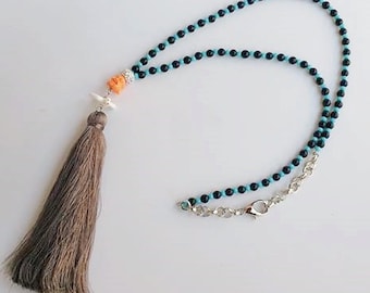 Puede incluir: Un collar de borla marrón con cuentas negras y turquesa. El collar tiene un cierre plateado y un pequeño dije naranja y blanco.