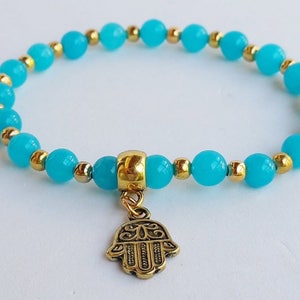 Pode incluir: Um bracelete de contas azul turquesa com um amuleto de mão de hamsa dourado.