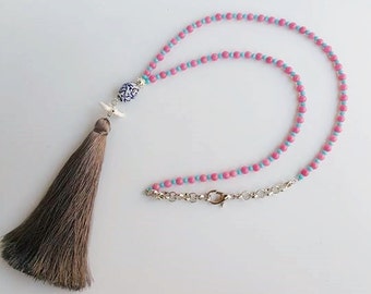 Puede incluir: Un collar de cuentas rosa y azul con un borla y un colgante de cerámica. El colgante tiene un diseño floral azul y blanco.