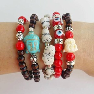 Pode incluir: Cinco pulseiras empilhadas em um pulso. As pulseiras são feitas de madeira, contas vermelhas, contas brancas e contas turquesa. Uma pulseira apresenta um amuleto de Buda turquesa, outra apresenta um amuleto de olho turco branco e outra apresenta um amuleto de Buda rindo branco.