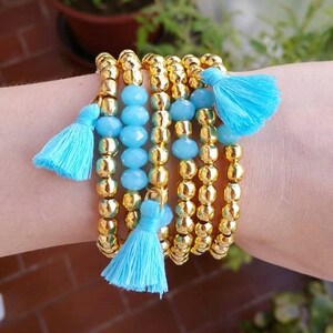 Pode incluir: Um conjunto de seis pulseiras com contas douradas e borlas azuis. As pulseiras estão empilhadas no pulso de uma pessoa.
