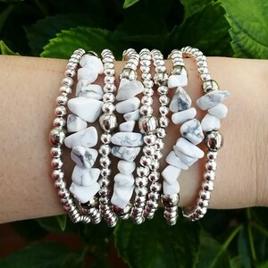 Pode incluir: Um conjunto de sete pulseiras com contas de pedra howlite branca e contas espaçadoras prateadas. As pulseiras estão empilhadas no pulso de uma pessoa.