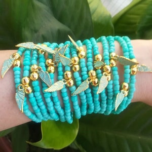 Pode incluir: Uma pilha de pulseiras de contas turquesa com pingentes de asas de anjo douradas.