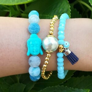 Pode incluir: Três pulseiras empilhadas em um pulso. A pulseira superior é feita de contas de ágata azul claro e branco com um amuleto de Buda turquesa. A pulseira do meio é feita de contas douradas. A pulseira inferior é feita de contas de vidro azul claro com um amuleto dourado e uma borla azul.