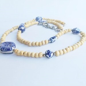 May include: A white and blue ceramic bead necklace with a wooden bead base. The necklace features a square ceramic bead with a blue and white design, a round ceramic bead with a blue and white floral design, and a round ceramic bead with a blue and white geometric design.