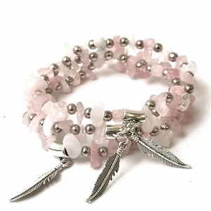 Puede incluir: Un brazalete de piedras preciosas rosas y blancas con cuentas plateadas y dijes de plumas.