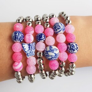 Pode incluir: Conjunto de cinco pulseiras de ágata rosa e branca com contas de metal prateado e pingentes de porcelana azul e branca. Os pingentes apresentam caracteres chineses, uma borboleta e um pássaro.