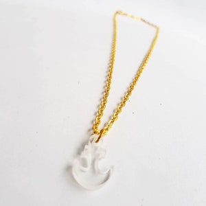 Pode incluir: Um colar de corrente dourada com um pingente de âncora de vidro transparente fosco. O colar é exibido em uma superfície branca, mostrando o design delicado da joia. O pingente de âncora é um símbolo de esperança e estabilidade.