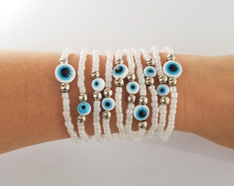 May include: White beaded bracelets with blue and white evil eye charms. The bracelets are made of small white beads and silver-colored metal beads. The evil eye charms have a blue iris and a black pupil.