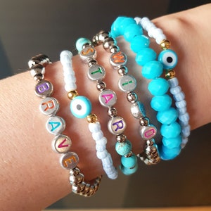 Pode incluir: Um conjunto de seis pulseiras com contas prateadas e pingentes de letras coloridas que soletram as palavras "BRAVE", "SMART", "WARD" e "WILD". Duas das pulseiras também têm um amuleto de olho turco azul.