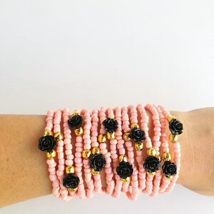 Op de afbeelding: Een stapel roze kralenarmbanden met zwarte roosaccenten. Elke armband heeft een gouden kraal tussen elke roos.