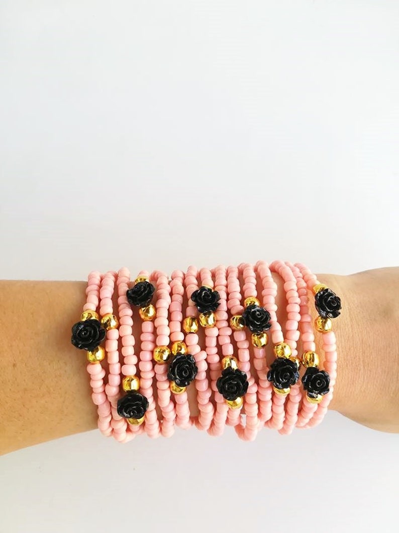 Op de afbeelding: Een stapel roze kralenarmbanden met zwarte roosaccenten. Elke armband heeft een gouden kraal tussen elke roos.