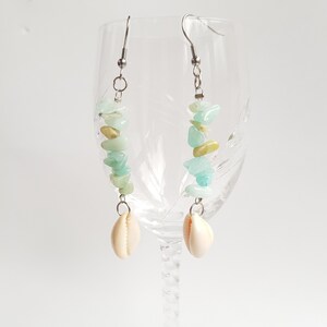 May include: A pair of dangle earrings with a silver hook and a chain. The earrings are made with light green and gold stones and a white seashell charm.