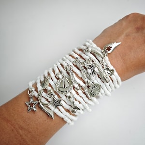 Pode incluir: Um conjunto de pulseiras de contas brancas com pingentes prateados, incluindo uma estrela, um veleiro, uma pena e um sol. As pulseiras são usadas no pulso.