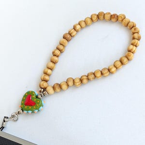 Pode incluir: Uma pulseira de contas de madeira com um pingente em forma de coração. O pingente é verde com um coração vermelho e detalhes em azul e laranja. A pulseira é feita de pequenas contas de madeira marrom claro.