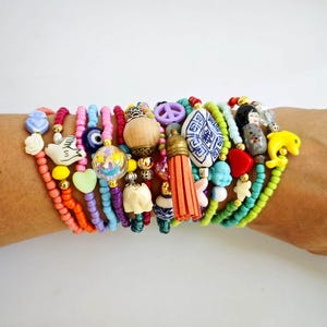 Pode incluir: Uma pilha de pulseiras de contas coloridas com vários pingentes, incluindo um sinal de paz, uma tartaruga, um mau-olhado, um coração, um pássaro, uma flor e um tassel.