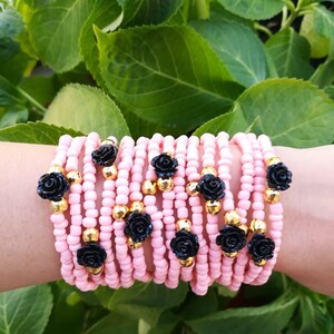 Op de afbeelding: Een stapel roze kralenarmbanden met zwarte roosaccenten en gouden kralen.
