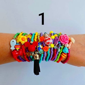 Pode incluir: Um conjunto de pulseiras de contas coloridas com vários encantos, incluindo uma cabeça do Mickey Mouse, uma flor, um peixe e um laço vermelho. As pulseiras estão no pulso de uma pessoa.