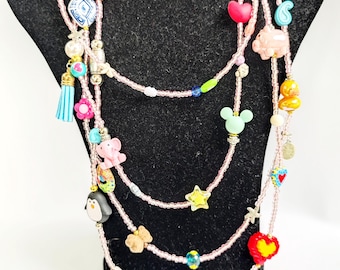 May include: A multi-strand necklace featuring light pink seed beads interspersed with a variety of colorful charms, including a blue tassel, a penguin, a pink elephant, a yellow star, a green mouse head silhouette, and various heart shapes.