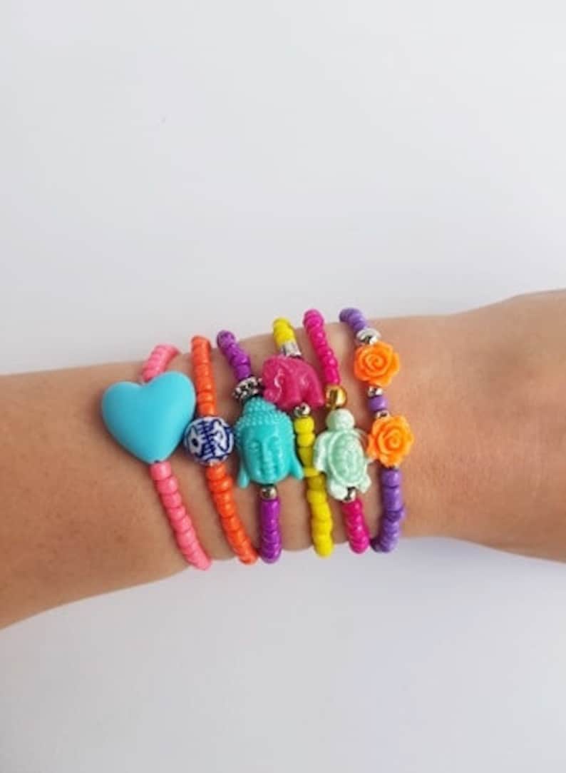 May include: A set of colourful beaded bracelets with various charms, including a heart, a Buddha head, a turtle, and a flower. The bracelets are stacked on a person's wrist.