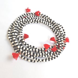 Pode incluir: Uma coleção de colares de contas com contas pretas e brancas alternadas. Cada colar é adornado com pequenas borlas vermelhas. Os colares estão dispostos em um padrão circular sobre um fundo branco.