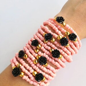 Op de afbeelding: Een stapel roze kralenarmbanden met zwarte roosaccenten en gouden kralen.