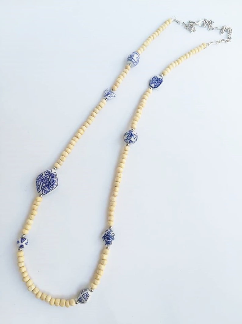May include: A long necklace made of light brown wooden beads with blue and white porcelain accents. The porcelain accents feature intricate designs.