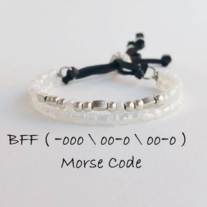 Pode incluir: Um bracelete de contas brancas com contas prateadas e um cordão preto. O bracelete tem o texto "BFF (-000 / 00-0 / 00-0)" e "Código Morse" escrito abaixo.