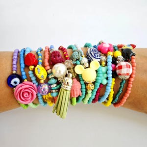 Pode incluir: Uma pilha de pulseiras de contas coloridas com vários pingentes, incluindo uma rosa rosa, um olho turco azul, uma cabeça de Mickey Mouse amarela, um coração vermelho, uma pérola branca, uma tartaruga turquesa, um Buda vermelho e um tassel verde.