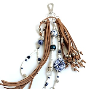Pode incluir: Um pendente decorativo para mala com borlas de camurça castanha, contas brancas e azul-escuras, detalhes em metal prateado e dois berloques em forma de concha e trevo de quatro folhas, preso a um mosquetão de metal prateado.