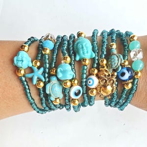 Pode incluir: Uma coleção de pulseiras de enrolar com contas teal. Cada pulseira apresenta detalhes dourados e amuletos, incluindo cabeças de Buda, estrelas do mar, tartarugas e uma coruja. Amuletos adicionais incluem contas azuis de mau-olhado e contas de cristal transparente.