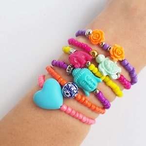 May include: A stack of colourful beaded bracelets with various charms, including a heart, a Buddha, a turtle, and flowers. The bracelets are on a person's wrist.