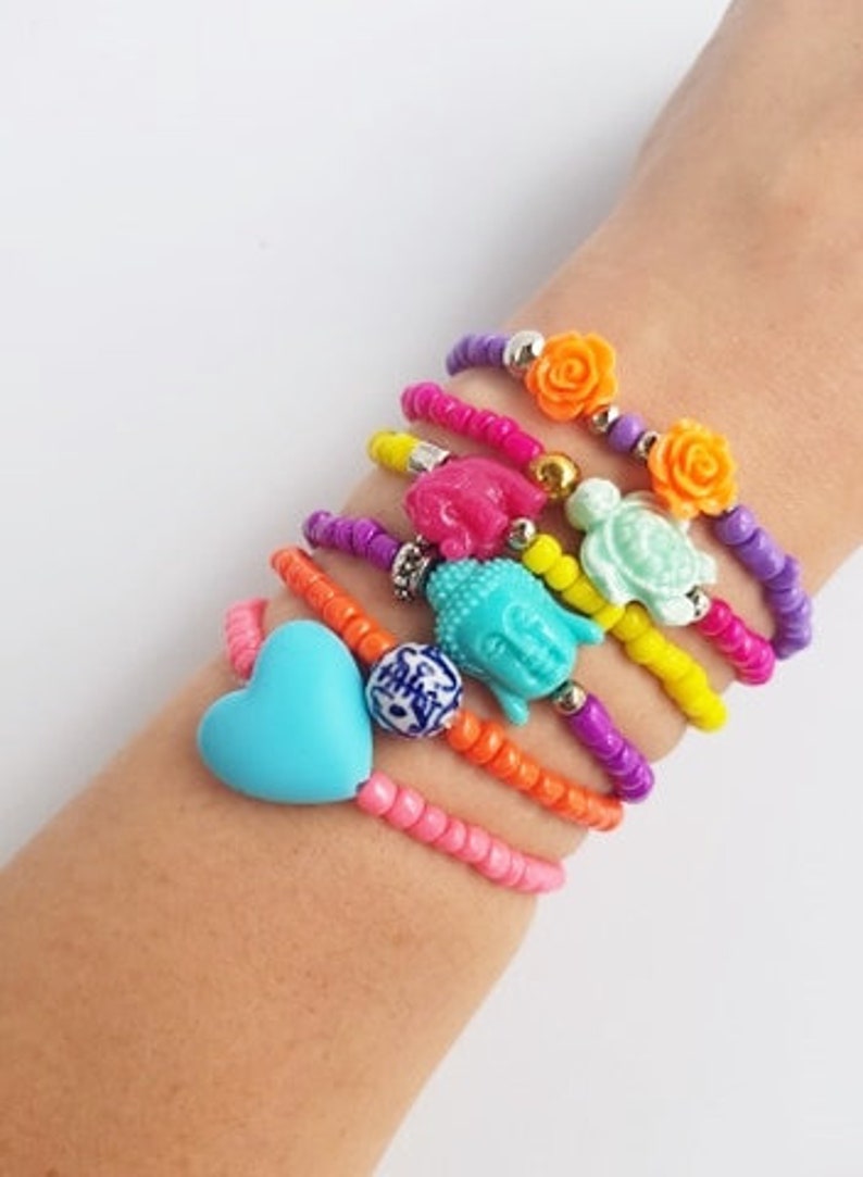 May include: A stack of colourful beaded bracelets with various charms, including a heart, a Buddha, a turtle, and flowers. The bracelets are on a person's wrist.