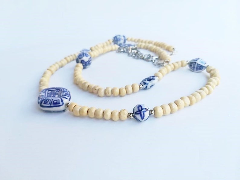 May include: A white and blue ceramic bead necklace with a wooden bead base. The necklace features a square ceramic bead with a blue and white design, a round ceramic bead with a blue and white floral design, and a round ceramic bead with a blue and white geometric design.